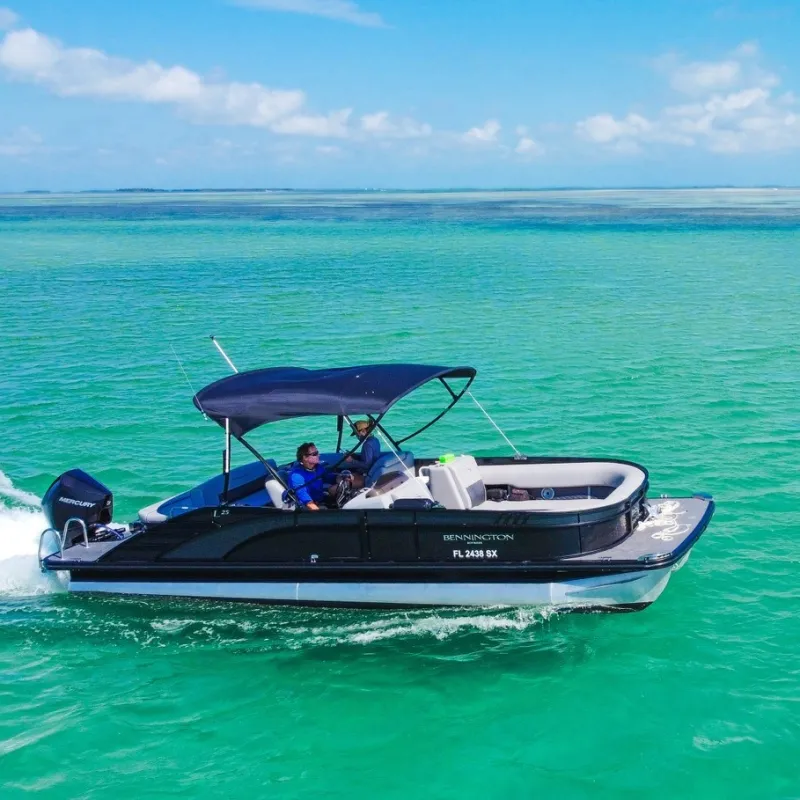 Two people on a black pontoon boat cruising in clear turquoise water under blue skies.