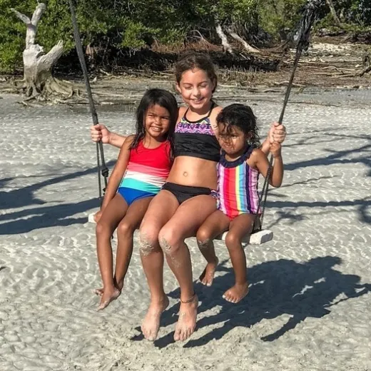 Three kids smiling, swinging on a beach, with trees in the background.