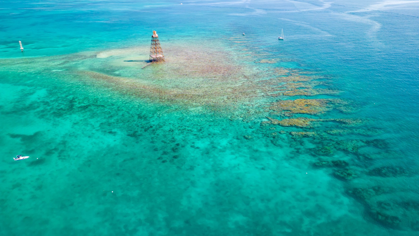 Aerial view of turquoise ocean with coral reefs, boats, and a metal tower.