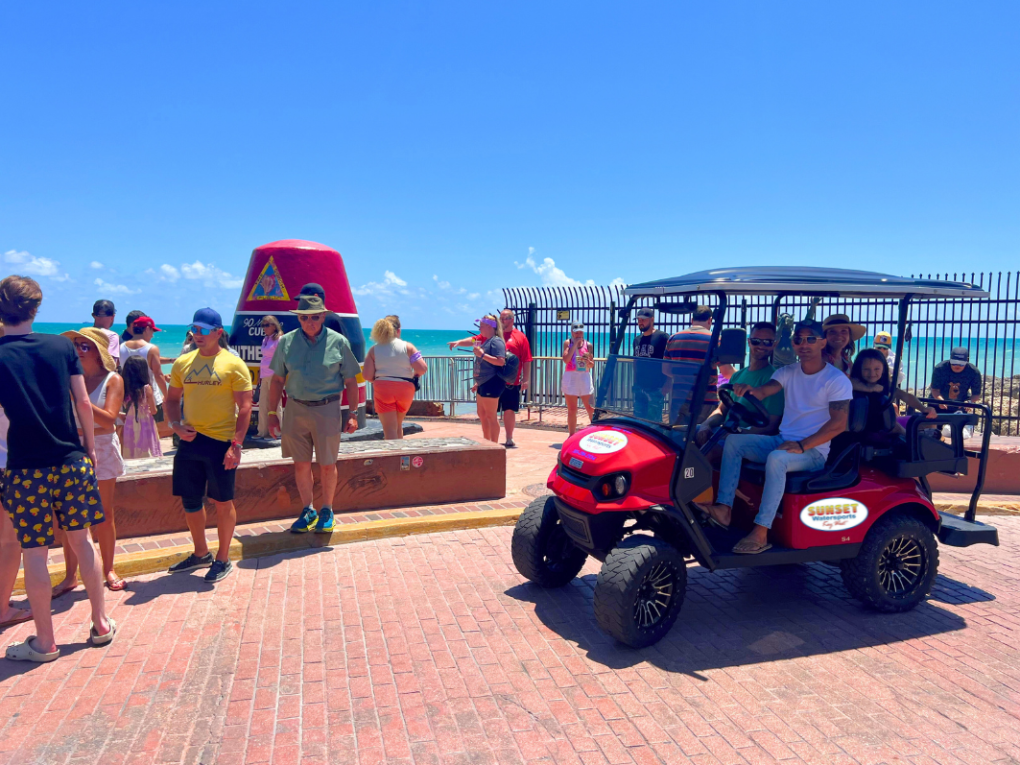 Tourists at Southernmost Point marker in Key West with red golf cart nearby.