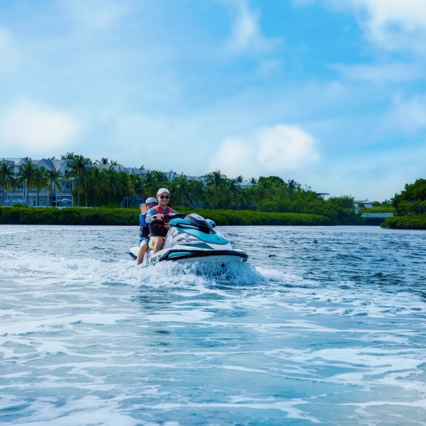 Two people riding a jet ski on a sunny day, with buildings and palm trees in the background.