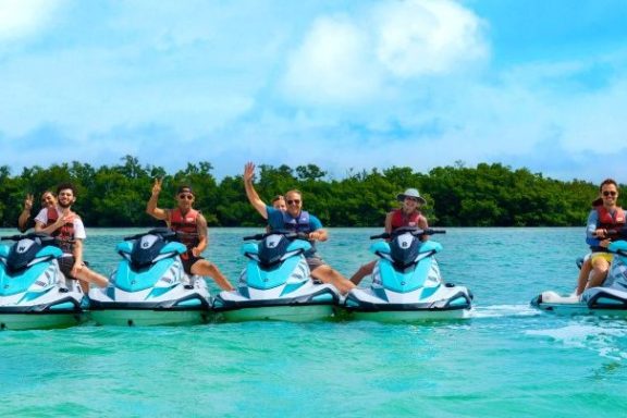 Five people on jet skis waving in turquoise water with trees and blue sky background.