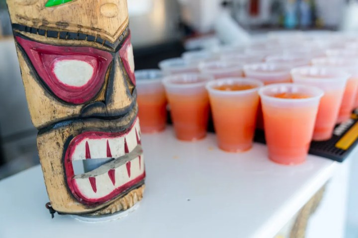 Tiki mask on a bar next to rows of orange drinks in plastic cups.