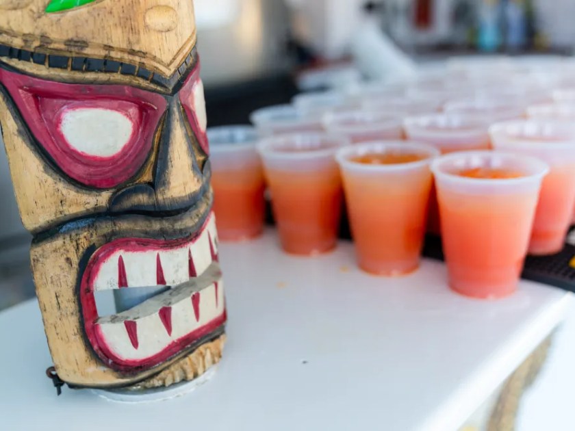 Tiki mask on a bar next to rows of orange drinks in plastic cups.
