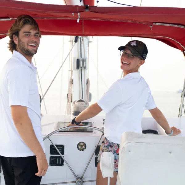 Two people smiling on a boat with a red canopy, one steering the wheel.