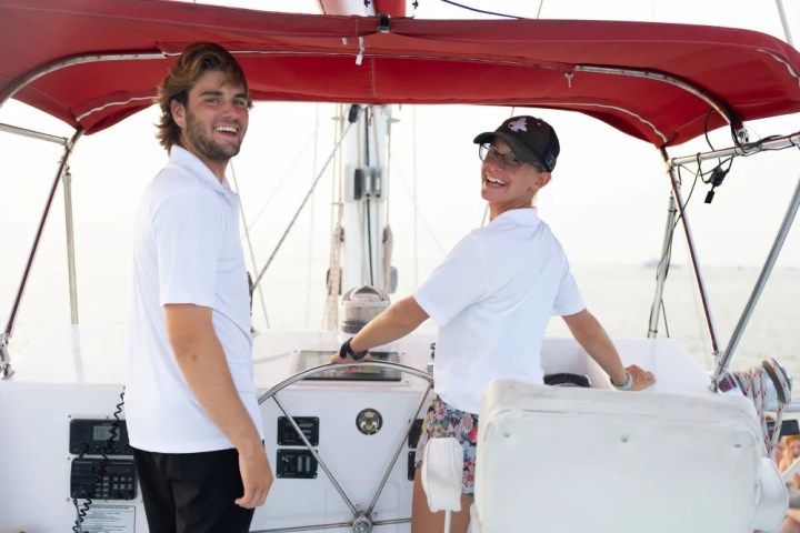 Two people smiling on a boat with a red canopy, one steering the wheel.